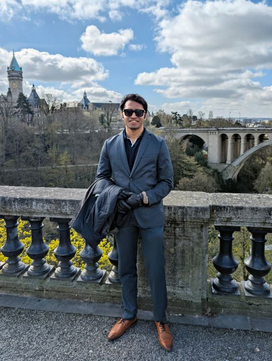 Manu Mythil Muralee standing at a stone balustrade in front of a stone arch bridge.